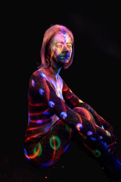 In a seated side pose, Selina's body is decorated with a colorful UV design combining orange and blue stripes with round dot patterns across the torso and legs. Bodypaint by Elad Itzkin. Photographed by Elad Itzkin Photography under UV light in his studio in Gesundbrunnen, Berlin.