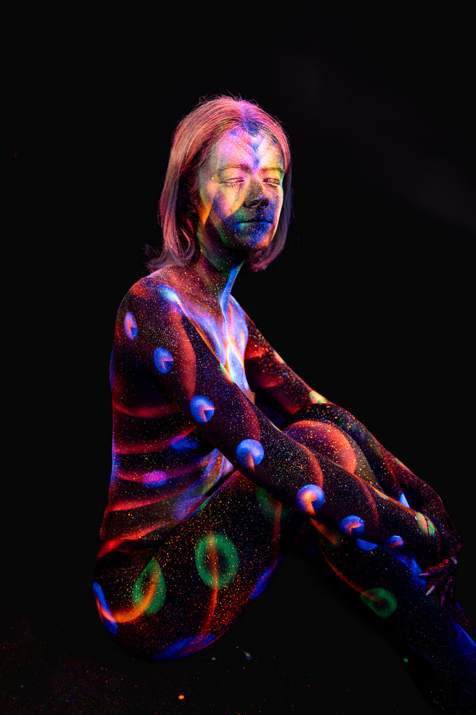 In a seated side pose, Selina's body is decorated with a colorful UV design combining orange and blue stripes with round dot patterns across the torso and legs. Bodypaint by Elad Itzkin. Photographed by Elad Itzkin Photography under UV light in his studio in Gesundbrunnen, Berlin.