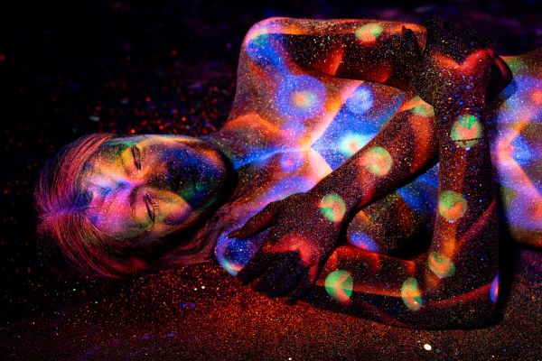 Selina sits with her body turned to the side, painted in a UV design of orange stripes and scattered dots across the torso and legs. Bodypaint by Elad Itzkin. Photographed by Elad Itzkin Photography under blacklight in his studio in Gesundbrunnen, Berlin.