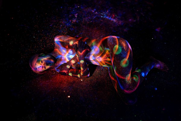 Selina lies curled on her side on the studio floor, her body covered in a vivid UV bodypaint design of swirling orange and blue linework with green dotted accents. Bodypaint by Elad Itzkin. Photographed by Elad Itzkin Photography under UV light in his studio in Gesundbrunnen, Berlin.