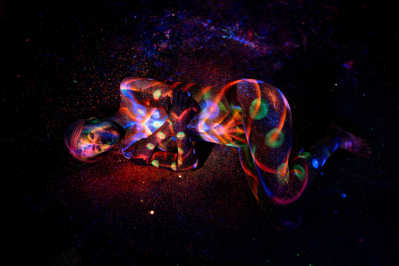 Selina lies curled on her side on the studio floor, her body covered in a vivid UV bodypaint design of swirling orange and blue linework with green dotted accents. Bodypaint by Elad Itzkin. Photographed by Elad Itzkin Photography under UV light in his studio in Gesundbrunnen, Berlin.