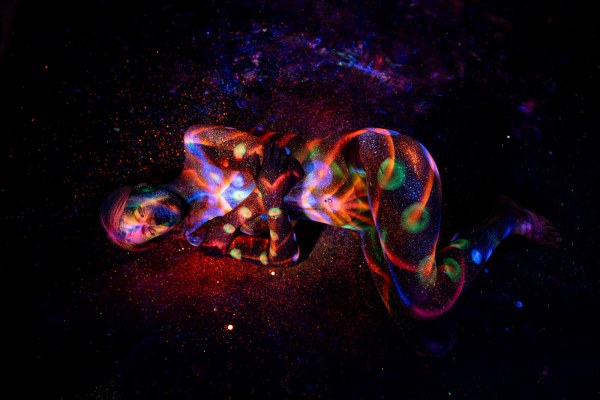 Lying on her side in a curled position, Selina's body is painted with a colorful UV pattern of swirling orange and blue linework accented with green dots. Bodypaint by Elad Itzkin. Photographed by Elad Itzkin Photography under blacklight in his studio in Gesundbrunnen, Berlin.