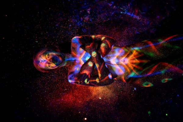 Selina lies in a curled position with her head tilted to one side, her torso and legs decorated with a bright UV design of orange and blue linework and green dotted accents. Bodypaint by Elad Itzkin. Photographed by Elad Itzkin Photography under UV light in his studio in Gesundbrunnen, Berlin.