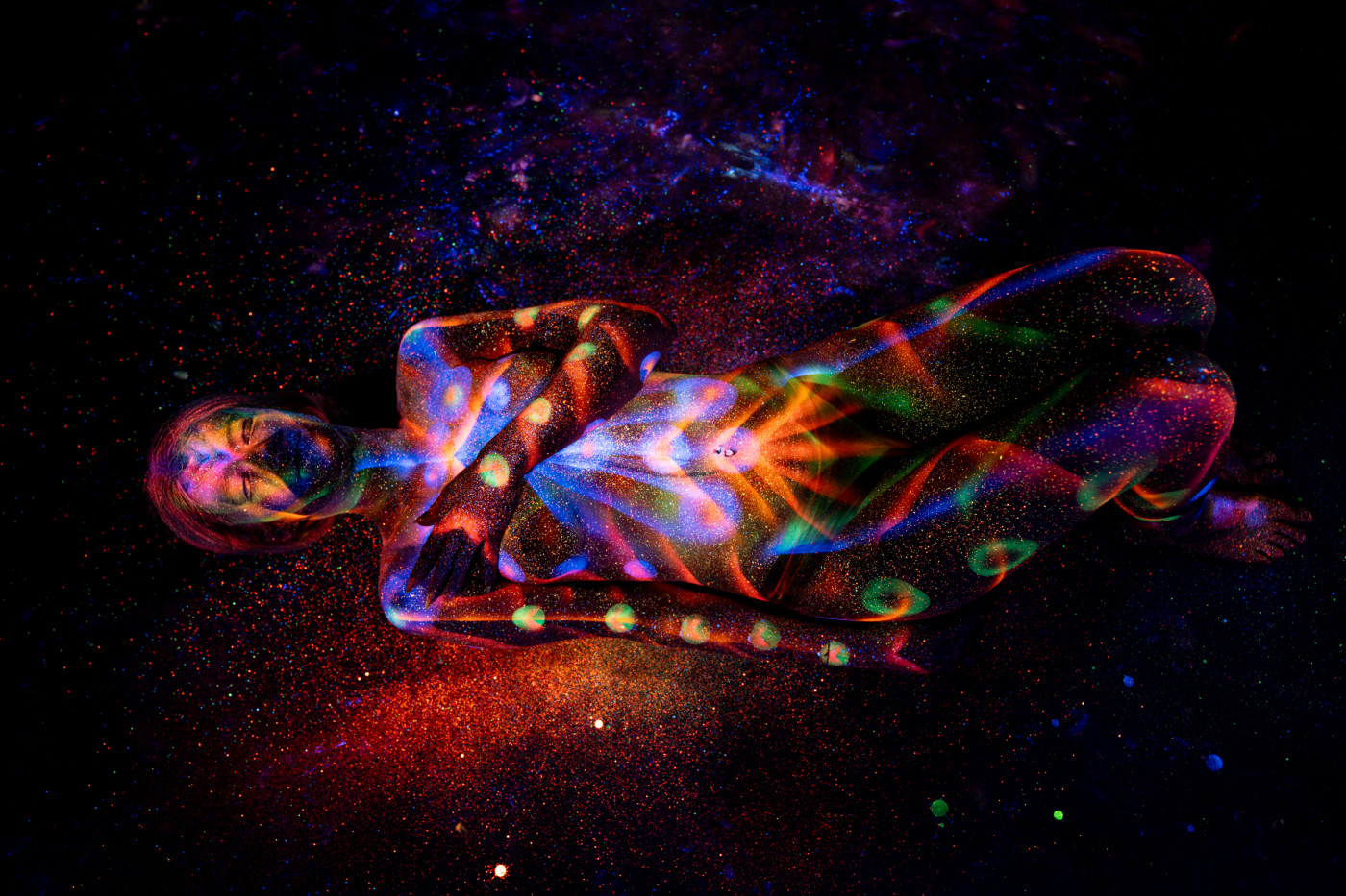 In a curled, reclining position, Selina's body is marked with a vivid UV bodypaint pattern of orange and blue swirls with scattered green dots. Bodypaint by Elad Itzkin. Photographed by Elad Itzkin Photography under blacklight in his studio in Gesundbrunnen, Berlin.