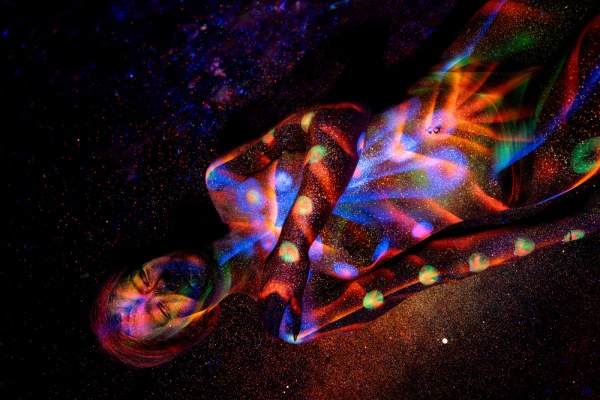 Model Selina lies on her side with her head tilted toward the camera. Her bodypaint combines orange, blue and green polka dots with radiating linework across her torso and legs. Bodypaint by Elad Itzkin. Photographed by Elad Itzkin Photography under UV/blacklight in his studio in Gesundbrunnen, Berlin.
