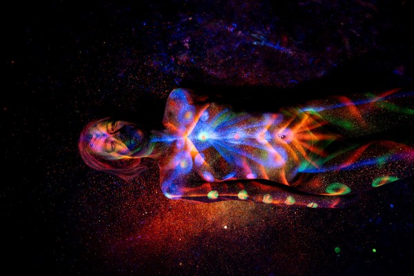 Selina lies on her back, photographed from above with her head turned toward the camera. A radiating orange and blue starburst pattern covers her chest, transitioning into a row of glowing polka dots along her legs. Bodypaint by Elad Itzkin. Photographed by Elad Itzkin Photography under UV/blacklight in his studio in Gesundbrunnen, Berlin.
