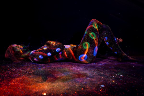 Selina lies on her side with her knees drawn up toward her chest. A repeating pattern of glowing blue, green and orange polka dots covers her body against the black background. Bodypaint by Elad Itzkin. Photographed by Elad Itzkin Photography under UV/blacklight in his studio in Gesundbrunnen, Berlin.