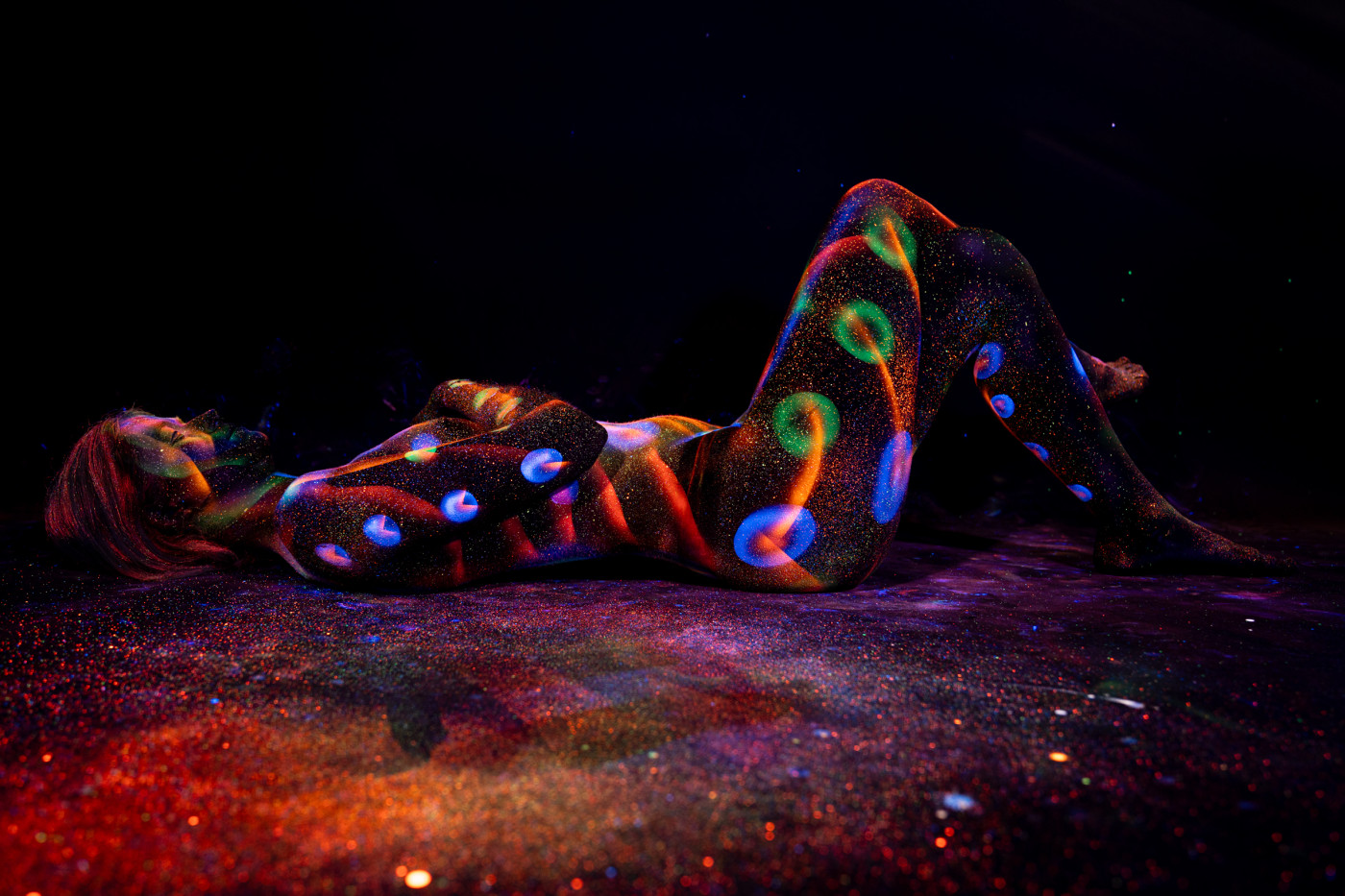 Selina lies on her side with her knees drawn up toward her chest. A repeating pattern of glowing blue, green and orange polka dots covers her body against the black background. Bodypaint by Elad Itzkin. Photographed by Elad Itzkin Photography under UV/blacklight in his studio in Gesundbrunnen, Berlin.