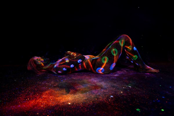 Lying on her back with her knees bent, Selina is painted with a polka dot design in blue, green and orange tones spanning her legs and torso. Bodypaint by Elad Itzkin. Photographed by Elad Itzkin Photography under UV/blacklight in his studio in Gesundbrunnen, Berlin.