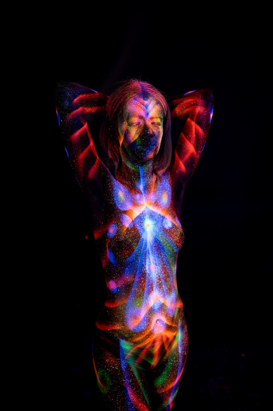 Selina stands facing the camera. Her bodypaint combines a symmetrical red flame-like pattern along the sides of her torso with a blue and white radiating starburst design across her chest and abdomen. Bodypaint by Elad Itzkin. Photographed by Elad Itzkin Photography under UV/blacklight in his studio in Gesundbrunnen, Berlin.