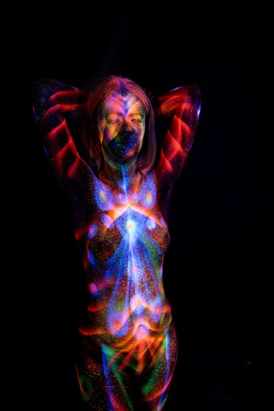Standing in a frontal pose, Selina wears a symmetrical bodypaint design that combines red radiating linework with a blue and orange starburst pattern centered on her chest. Bodypaint by Elad Itzkin. Photographed by Elad Itzkin Photography under UV/blacklight in his studio in Gesundbrunnen, Berlin.