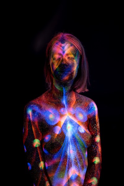 Selina stands facing forward. Her torso is covered in a symmetrical design of blue, orange and green tones, with rows of glowing polka dots running down each side. Bodypaint by Elad Itzkin. Photographed by Elad Itzkin Photography under UV/blacklight in his studio in Gesundbrunnen, Berlin.