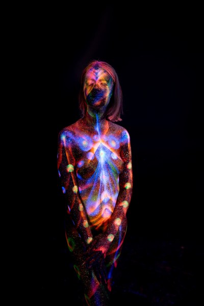 In a standing frontal pose, Selina's bodypaint shows a symmetrical radiating pattern in blue and orange across her chest, bordered by a line of glowing polka dots down each side of her torso. Bodypaint by Elad Itzkin. Photographed by Elad Itzkin Photography under UV/blacklight in his studio in Gesundbrunnen, Berlin.