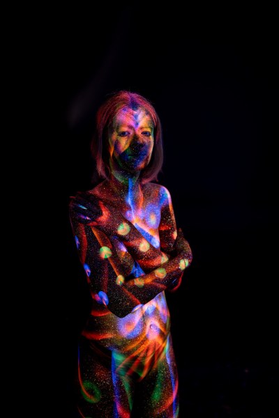Selina stands in a frontal pose facing the camera. A geometric bodypaint design in blue, orange and green covers her torso, accented with clusters of glowing polka dots along her side. Bodypaint by Elad Itzkin. Photographed by Elad Itzkin Photography under UV/blacklight in his studio in Gesundbrunnen, Berlin.