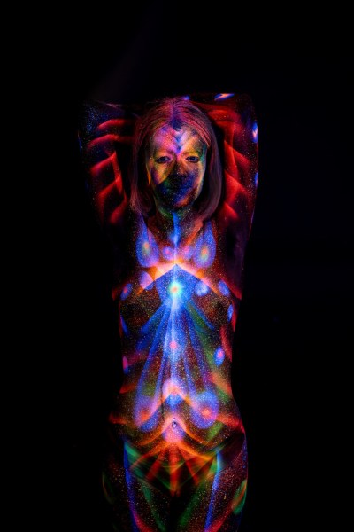 Facing the camera in a standing pose, Selina is painted with a symmetrical design of red radiating lines and a blue and orange starburst pattern centered on her torso. Bodypaint by Elad Itzkin. Photographed by Elad Itzkin Photography under UV/blacklight in his studio in Gesundbrunnen, Berlin.