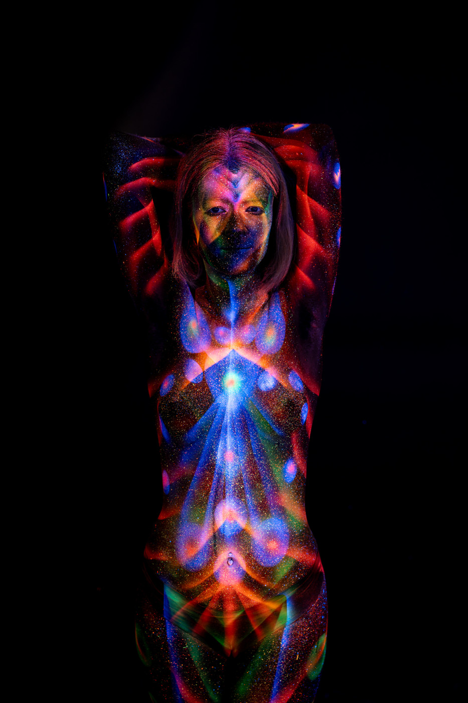 Facing the camera in a standing pose, Selina is painted with a symmetrical design of red radiating lines and a blue and orange starburst pattern centered on her torso. Bodypaint by Elad Itzkin. Photographed by Elad Itzkin Photography under UV/blacklight in his studio in Gesundbrunnen, Berlin.