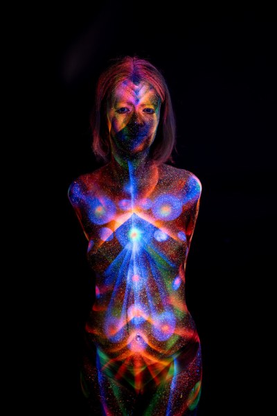 Selina stands facing forward, her torso covered in a symmetrical bodypaint pattern of blue, orange and green tones radiating outward from the center of her chest. Bodypaint by Elad Itzkin. Photographed by Elad Itzkin Photography under UV/blacklight in his studio in Gesundbrunnen, Berlin.
