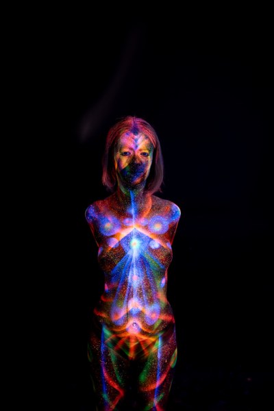 In a frontal standing pose, Selina's bodypaint design features a symmetrical arrangement of blue and orange radiating lines across her chest and abdomen. Bodypaint by Elad Itzkin. Photographed by Elad Itzkin Photography under UV/blacklight in his studio in Gesundbrunnen, Berlin.