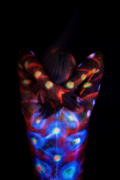 Selina stands with her back to the camera. Rows of glowing orange and green polka dots run along her sides, while a blue floral pattern spreads across her upper back. Bodypaint by Elad Itzkin. Photographed by Elad Itzkin Photography under UV/blacklight in his studio in Gesundbrunnen, Berlin.