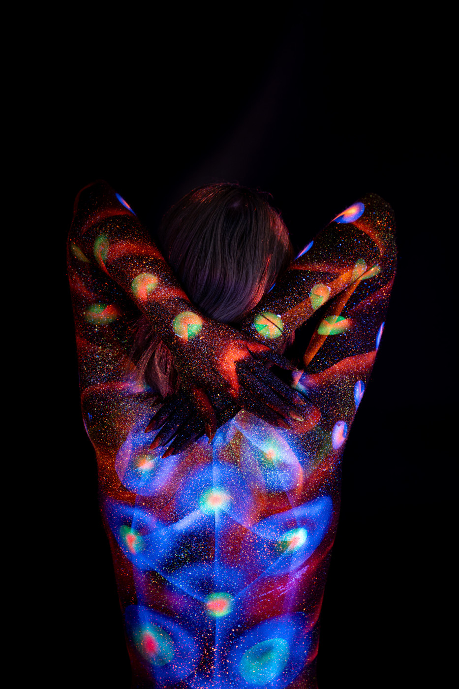 Selina stands with her back to the camera. Rows of glowing orange and green polka dots run along her sides, while a blue floral pattern spreads across her upper back. Bodypaint by Elad Itzkin. Photographed by Elad Itzkin Photography under UV/blacklight in his studio in Gesundbrunnen, Berlin.