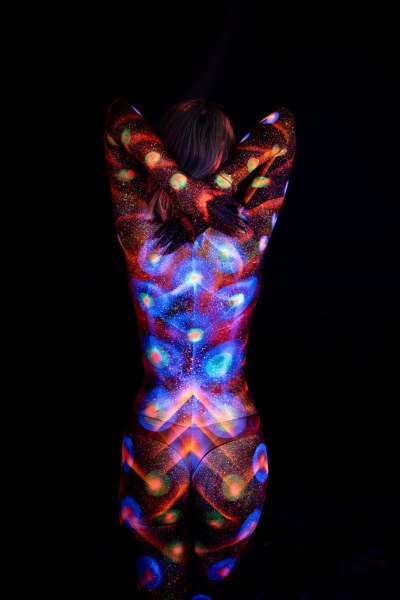 With her back to the camera, Selina's bodypaint combines glowing polka dots along her sides with a symmetrical blue and orange pattern radiating across her back and lower body. Bodypaint by Elad Itzkin. Photographed by Elad Itzkin Photography under UV/blacklight in his studio in Gesundbrunnen, Berlin.