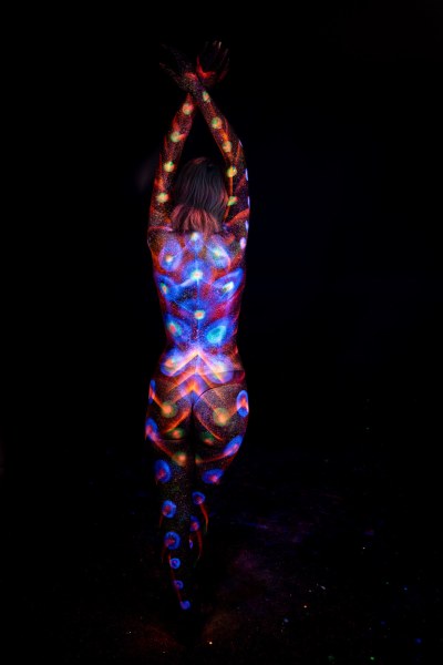 Standing with her back to the camera, Selina's bodypaint features a chain of glowing dots in orange and green tones alongside a symmetrical blue and purple pattern radiating across her back. Bodypaint by Elad Itzkin. Photographed by Elad Itzkin Photography under UV/blacklight in his studio in Gesundbrunnen, Berlin.