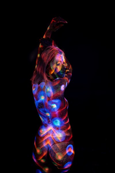 Selina is photographed from behind in a three-quarter turn. A symmetrical bodypaint pattern in blue and orange spans her back, bordered by rows of glowing polka dots along her side. Bodypaint by Elad Itzkin. Photographed by Elad Itzkin Photography under UV/blacklight in his studio in Gesundbrunnen, Berlin.