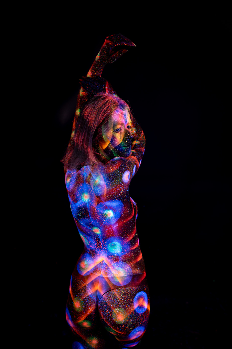 Selina is photographed from behind in a three-quarter turn. A symmetrical bodypaint pattern in blue and orange spans her back, bordered by rows of glowing polka dots along her side. Bodypaint by Elad Itzkin. Photographed by Elad Itzkin Photography under UV/blacklight in his studio in Gesundbrunnen, Berlin.