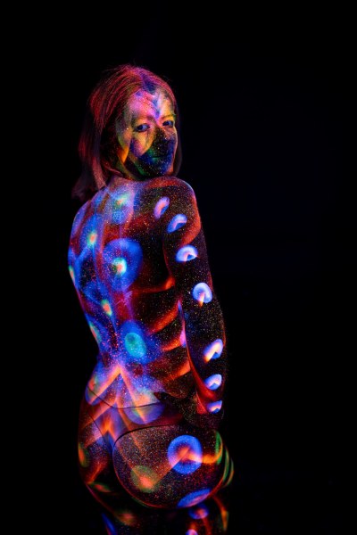 Selina kneels with her back to the camera, her head turned to look back over her shoulder. Her back and legs are covered in a repeating pattern of blue and orange polka dots. Bodypaint by Elad Itzkin. Photographed by Elad Itzkin Photography under UV/blacklight in his studio in Gesundbrunnen, Berlin.