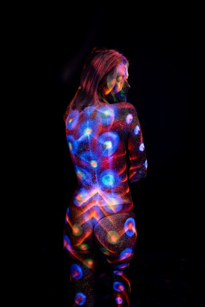 Kneeling with her back to the camera and her head turned toward the lens, Selina's bodypaint shows a symmetrical design of blue and orange polka dots and radiating linework across her back. Bodypaint by Elad Itzkin. Photographed by Elad Itzkin Photography under UV/blacklight in his studio in Gesundbrunnen, Berlin.