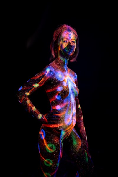 Selina stands in a three-quarter pose facing the camera. Her bodypaint combines glowing polka dots and radiating linework across her torso, with a circular linework design on one leg. Bodypaint by Elad Itzkin. Photographed by Elad Itzkin Photography under UV/blacklight in his studio in Gesundbrunnen, Berlin.