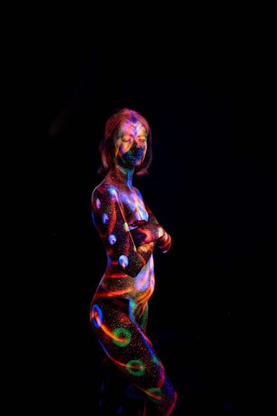 Selina stands in profile against a black background, her torso and legs marked with a UV reactive bodypaint design of red, blue, purple and green circular dots and a spiral swirl motif. Bodypaint by Elad Itzkin. Photographed by Elad Itzkin Photography under UV blacklight in his studio in Gesundbrunnen, Berlin.