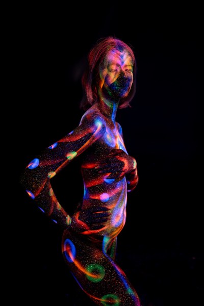 Selina stands in a three-quarter pose against a black background. A pattern of blue, red, purple and green circular dots and a spiral swirl covers her torso and hip in UV reactive bodypaint. Bodypaint by Elad Itzkin. Photographed by Elad Itzkin Photography under UV blacklight in his studio in Gesundbrunnen, Berlin.