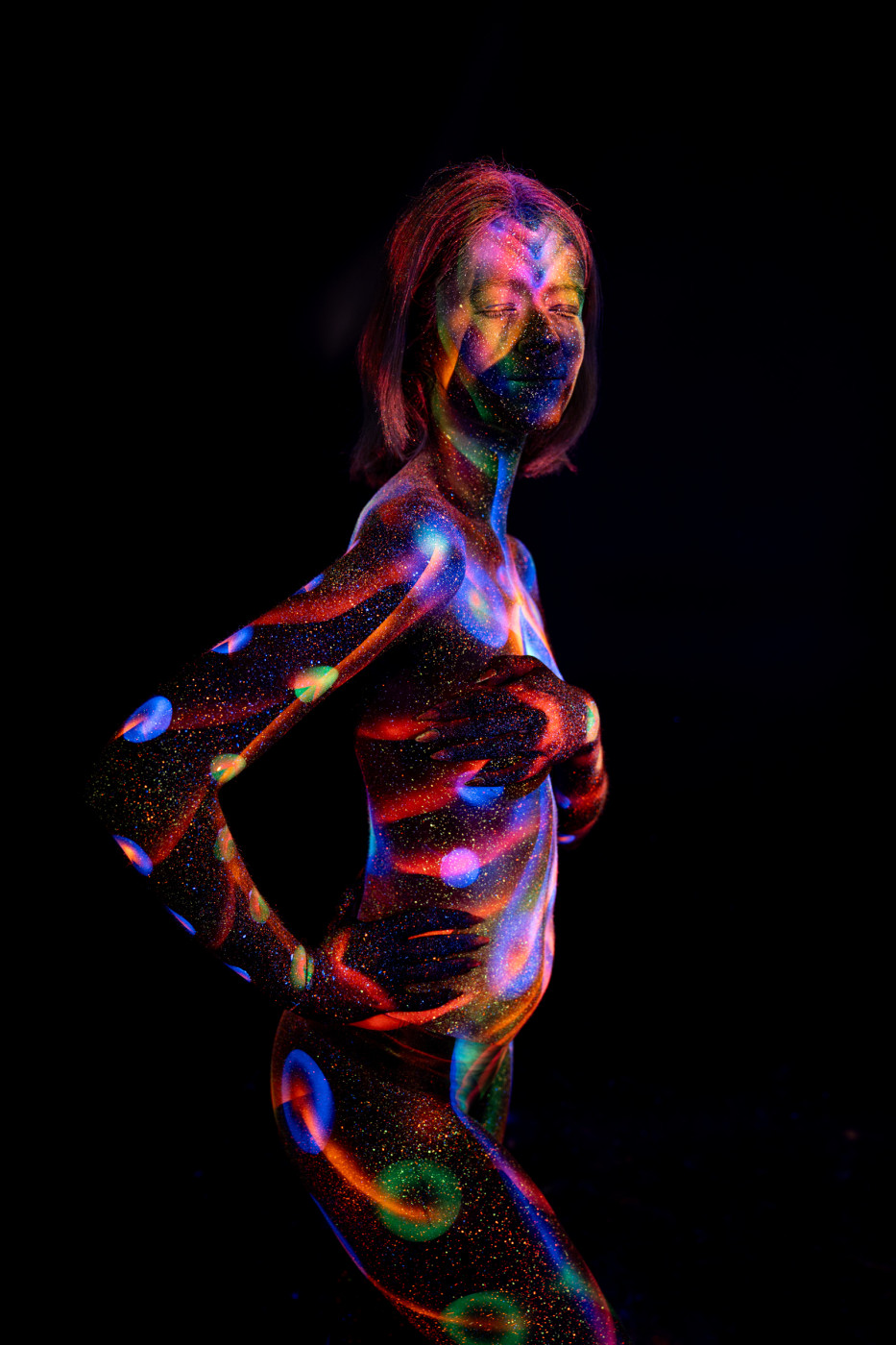 Selina stands in a three-quarter pose against a black background. A pattern of blue, red, purple and green circular dots and a spiral swirl covers her torso and hip in UV reactive bodypaint. Bodypaint by Elad Itzkin. Photographed by Elad Itzkin Photography under UV blacklight in his studio in Gesundbrunnen, Berlin.