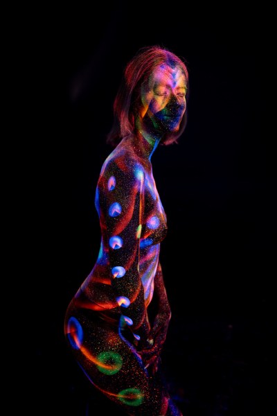 Selina stands in profile, her torso marked with a vertical row of circular UV reactive bodypaint shapes in blue, red and green, with a spiral swirl design near the hip. Bodypaint by Elad Itzkin. Photographed by Elad Itzkin Photography under UV blacklight in his studio in Gesundbrunnen, Berlin.
