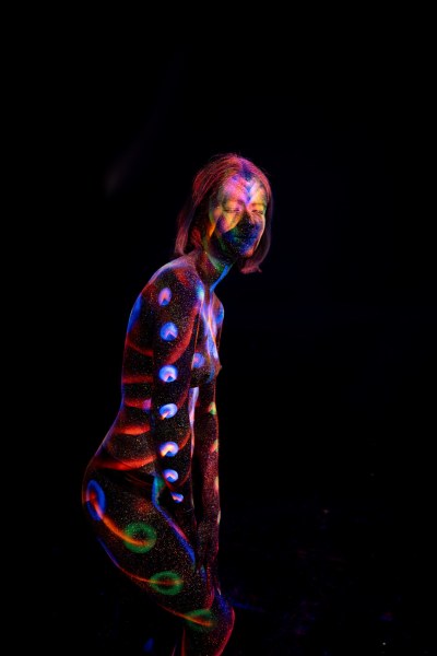 Standing in a three-quarter pose with a slight forward lean, Selina wears a UV reactive bodypaint design of red, blue and green circular dots arranged vertically down her torso and leg, with a spiral swirl accent. Bodypaint by Elad Itzkin. Photographed by Elad Itzkin Photography under UV blacklight in his studio in Gesundbrunnen, Berlin.