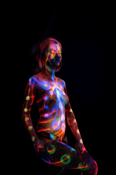 Selina stands in a three-quarter pose against a black background. Her body is painted with radiating UV reactive linework across the chest, circular dots along the torso, and a spiral swirl design at the hip, in red, blue, purple and green. Bodypaint by Elad Itzkin. Photographed by Elad Itzkin Photography under UV blacklight in his studio in Gesundbrunnen, Berlin.