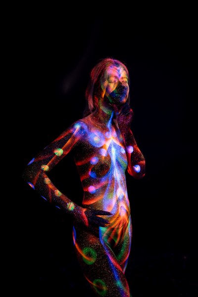 Selina stands in a three-quarter pose, her torso decorated with a UV reactive bodypaint pattern of circular dots, linework and a spiral swirl at the hip, in shades of red, blue, purple and green. Bodypaint by Elad Itzkin. Photographed by Elad Itzkin Photography under UV blacklight in his studio in Gesundbrunnen, Berlin.
