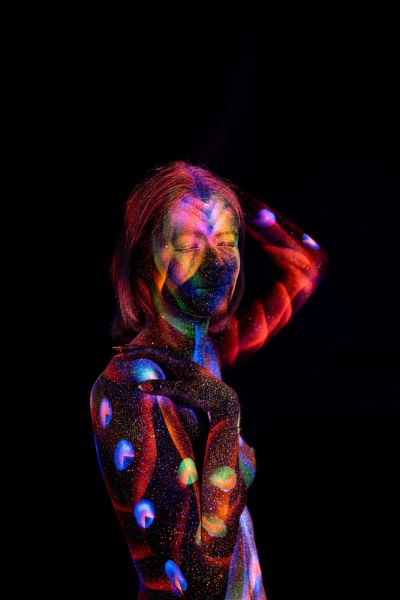 Selina stands in a three-quarter pose with her head turned, her torso covered in a UV reactive bodypaint pattern of circular dots and linework in blue, red, purple and green. Bodypaint by Elad Itzkin. Photographed by Elad Itzkin Photography under UV blacklight in his studio in Gesundbrunnen, Berlin.