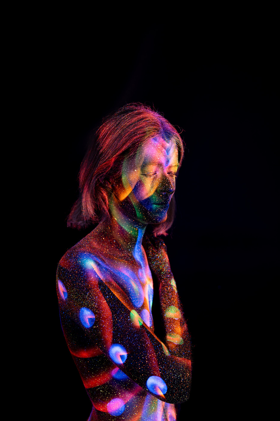 Selina stands in profile with her head tilted down. A UV reactive bodypaint design of circular dots and a spiral swirl in red, blue and green runs vertically down her torso and leg. Bodypaint by Elad Itzkin. Photographed by Elad Itzkin Photography under UV blacklight in his studio in Gesundbrunnen, Berlin.