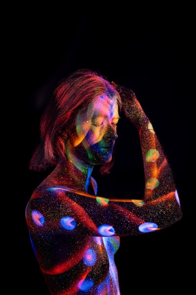 Selina stands in a three-quarter pose against a black background, her torso covered in a UV reactive bodypaint pattern of circular dots and linework in red, blue, purple and green. Bodypaint by Elad Itzkin. Photographed by Elad Itzkin Photography under UV blacklight in his studio in Gesundbrunnen, Berlin.