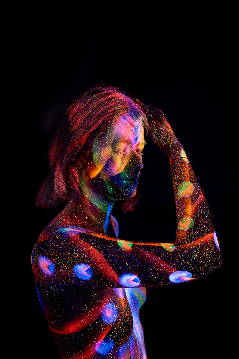 Selina stands in a three-quarter pose against a black background, her torso covered in a UV reactive bodypaint pattern of circular dots and linework in red, blue, purple and green. Bodypaint by Elad Itzkin. Photographed by Elad Itzkin Photography under UV blacklight in his studio in Gesundbrunnen, Berlin.