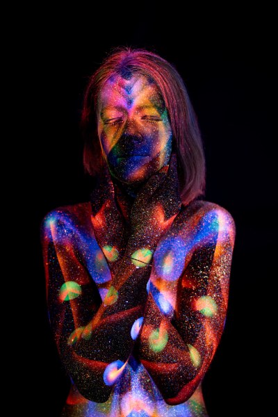 Selina stands facing forward, her chest and torso covered in a symmetrical UV reactive bodypaint design of chevron shapes and circular dots in blue, red, purple and green. Bodypaint by Elad Itzkin. Photographed by Elad Itzkin Photography under UV blacklight in his studio in Gesundbrunnen, Berlin.