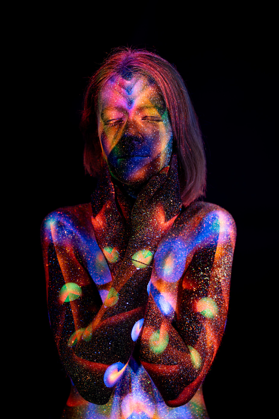 Selina stands facing forward, her chest and torso covered in a symmetrical UV reactive bodypaint design of chevron shapes and circular dots in blue, red, purple and green. Bodypaint by Elad Itzkin. Photographed by Elad Itzkin Photography under UV blacklight in his studio in Gesundbrunnen, Berlin.