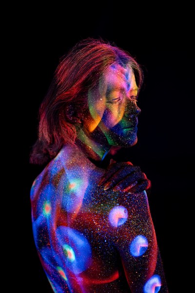 Selina stands with her back turned in a three-quarter pose, her shoulder and back covered in a UV reactive bodypaint pattern of circular dots in blue, purple and orange. Bodypaint by Elad Itzkin. Photographed by Elad Itzkin Photography under UV blacklight in his studio in Gesundbrunnen, Berlin.