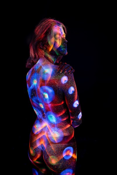 With her back turned in a three-quarter pose, Selina's back is marked with a UV reactive bodypaint design of chevron and zigzag linework with circular dot accents, in blue, red and orange. Bodypaint by Elad Itzkin. Photographed by Elad Itzkin Photography under UV blacklight in his studio in Gesundbrunnen, Berlin.