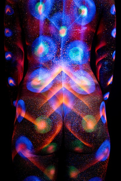 This image shows Selina's back and torso, turned away from the camera, marked with a symmetrical UV reactive bodypaint design of concentric circles and chevron shapes down the spine, in blue, teal, orange and pink. Bodypaint by Elad Itzkin. Photographed by Elad Itzkin Photography under UV blacklight in his studio in Gesundbrunnen, Berlin.