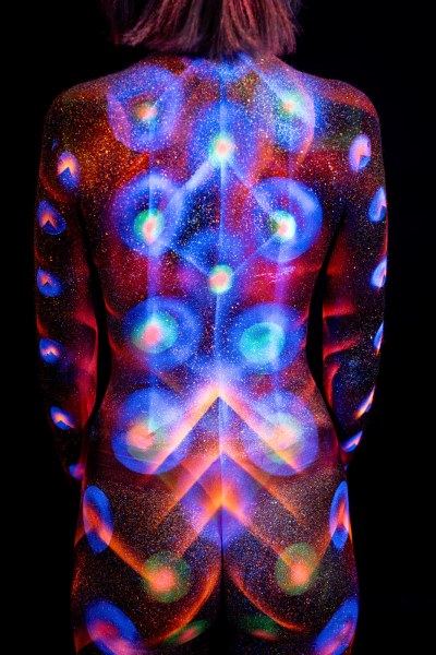 Selina stands with her back to the camera, head tilted down, her back covered in a symmetrical UV reactive bodypaint design of concentric circles and chevron shapes in blue, orange and pink. Bodypaint by Elad Itzkin. Photographed by Elad Itzkin Photography under UV blacklight in his studio in Gesundbrunnen, Berlin.
