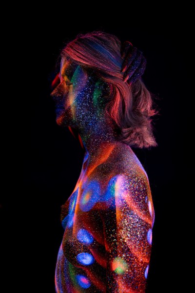 Selina stands in profile with her hair swept to one side, her torso marked with a vertical row of UV reactive bodypaint dots in blue, red and green. Bodypaint by Elad Itzkin. Photographed by Elad Itzkin Photography under UV blacklight in his studio in Gesundbrunnen, Berlin.
