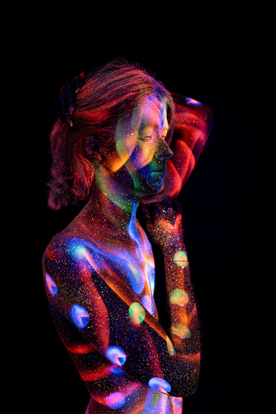 Selina stands in a three-quarter pose with her head tilted, her torso covered in a UV reactive bodypaint pattern of circular dots in blue, red, purple and green. Bodypaint by Elad Itzkin. Photographed by Elad Itzkin Photography under UV blacklight in his studio in Gesundbrunnen, Berlin.